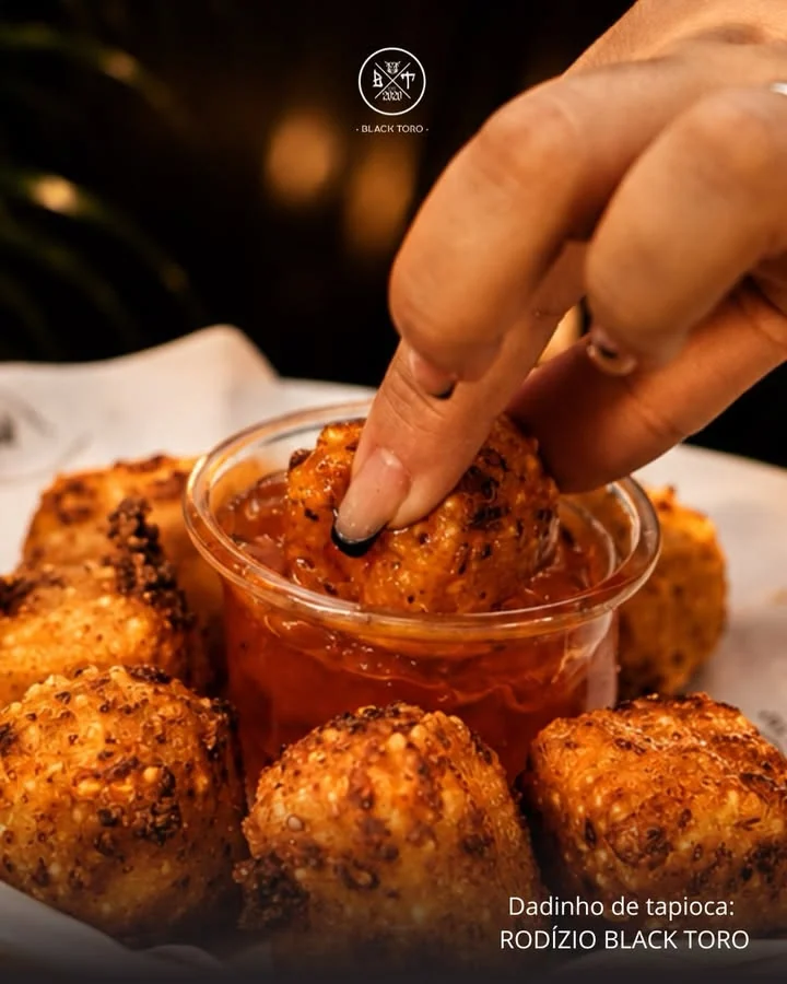 Dadinho de tapioca mergulhado no molho, com o brasão Black Toro e a legenda Rodízio Black Toro.